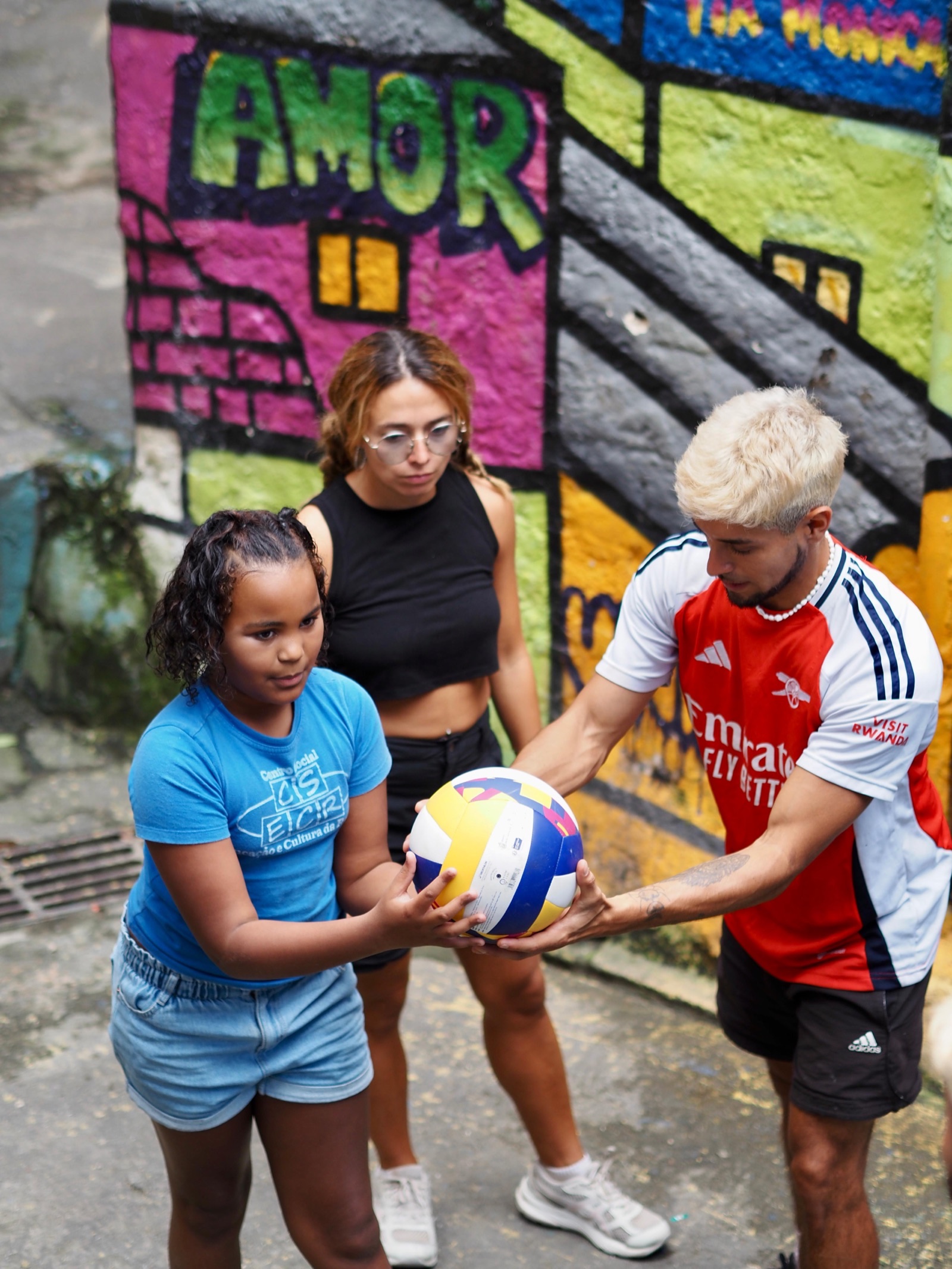 A volunteer and a woman hold a volleyball with a girl in front of a painted mural.