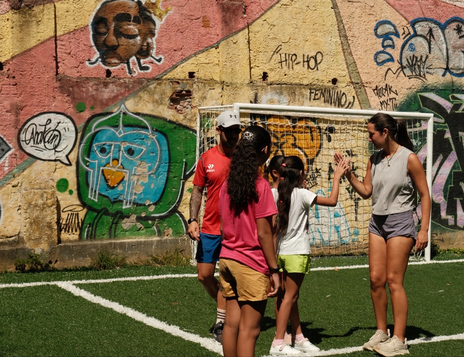 A volunteer high-fives children on the artificial pitch in Vidigal, in front of a goal and a wall covered in graffiti.