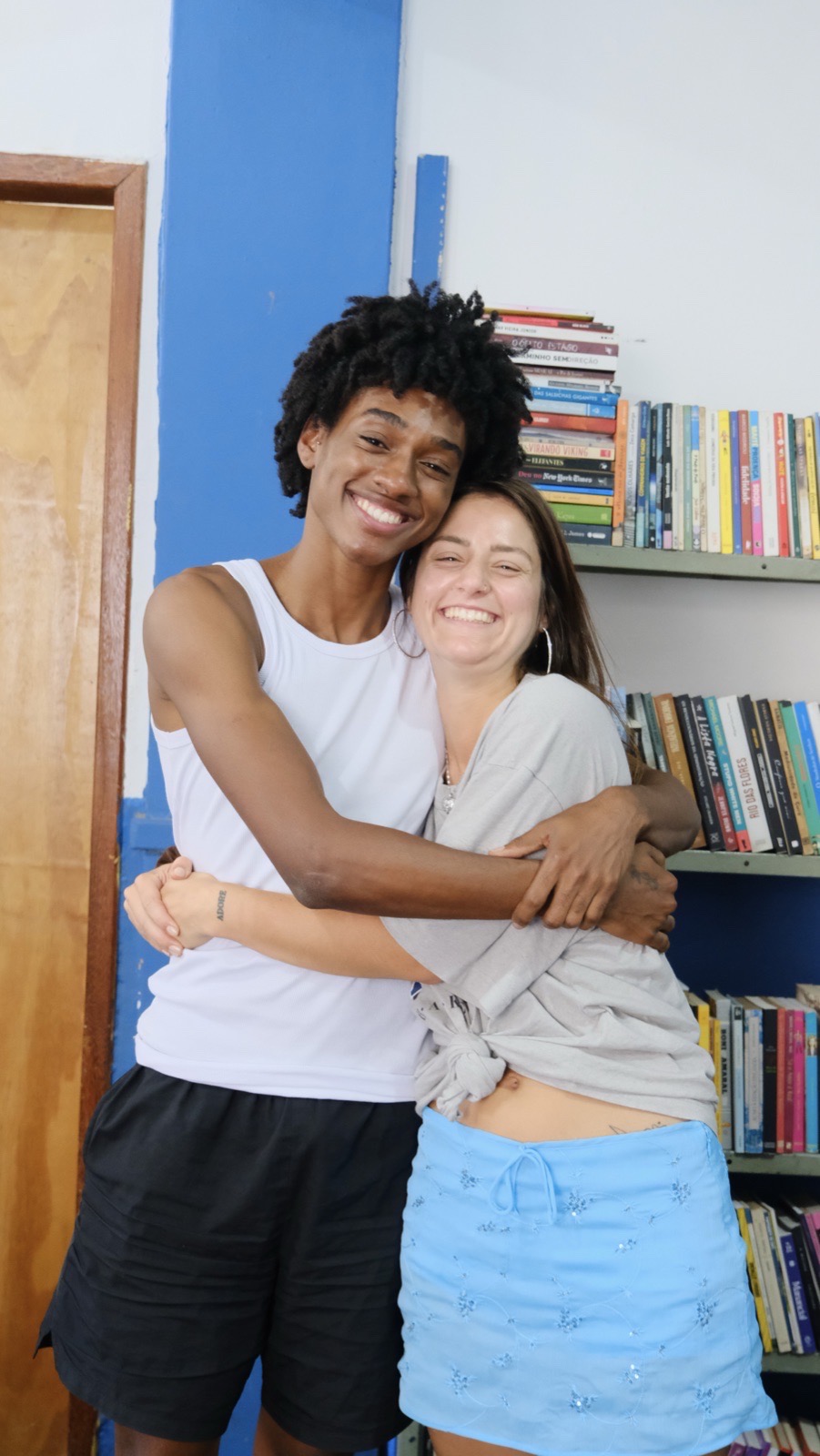 Two friends hug and smile beside a bookshelf in a community library.