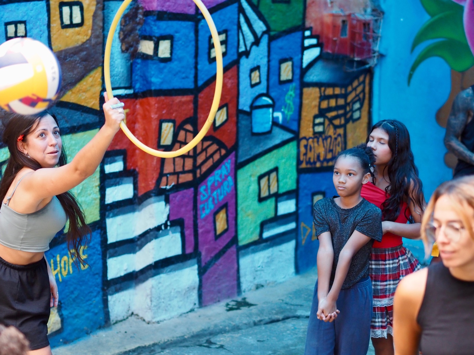 A volunteer holds up a hoop for a volleyball game while girls wait their turn in front of a colourful mural of houses.