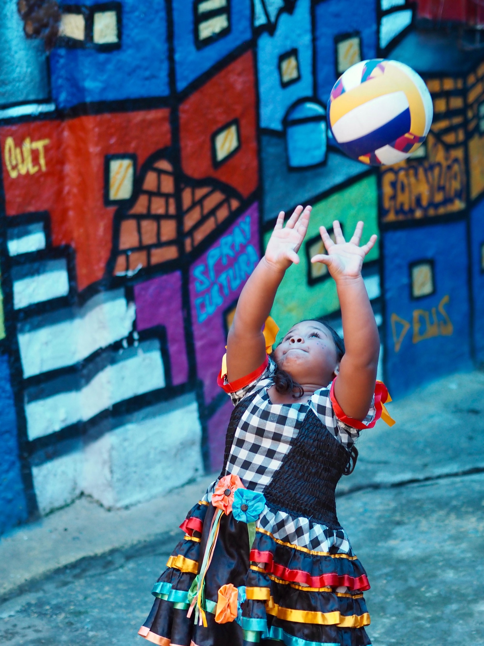 A girl in a checked, ruffled dress jumps to hit a volleyball in front of a brightly painted mural.