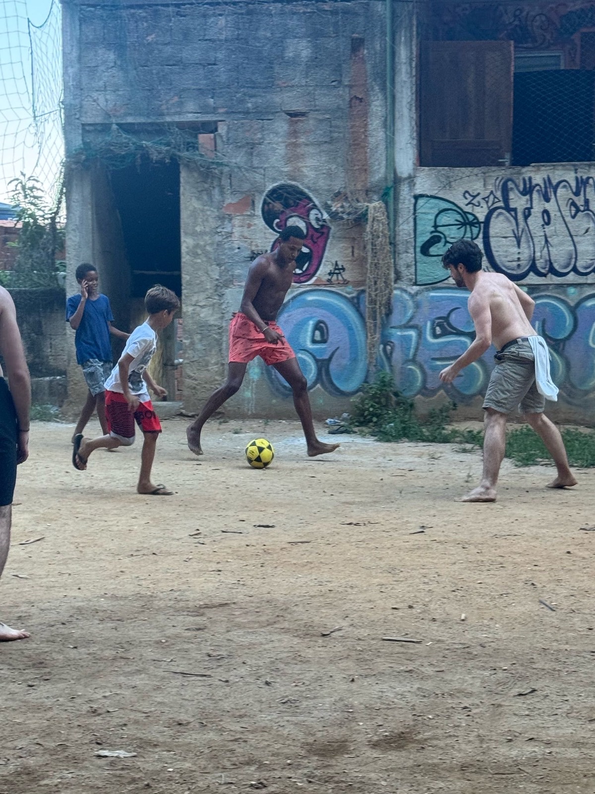 Children and a volunteer chasing a football across a dusty street pitch.