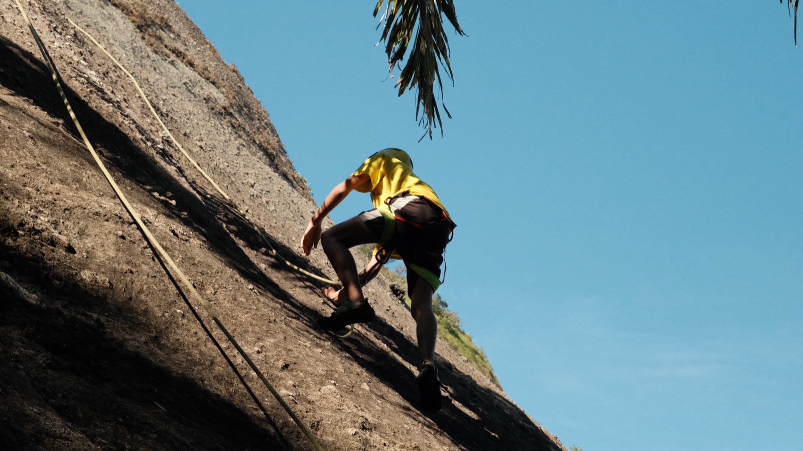 A climber in a yellow T-shirt scales a steep granite slope under a blue sky.